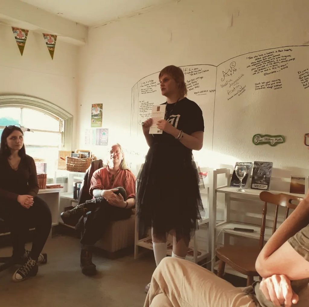 Laura Jane Round at the book launch of her second poetry pamphlet, TEATH. They are wearing a Nine Inch Nails shirt and tulle skirt. A crowd sits in a circle surrounding her.