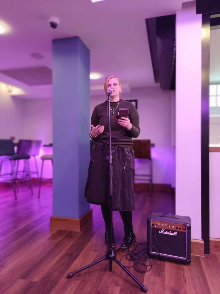 Laura Jane performs in-front of a microphone for the Saturday Books Gallery monthly Poetry Breakfast. The background has a purple hue. Round is dressed in green complete with green lipstick.